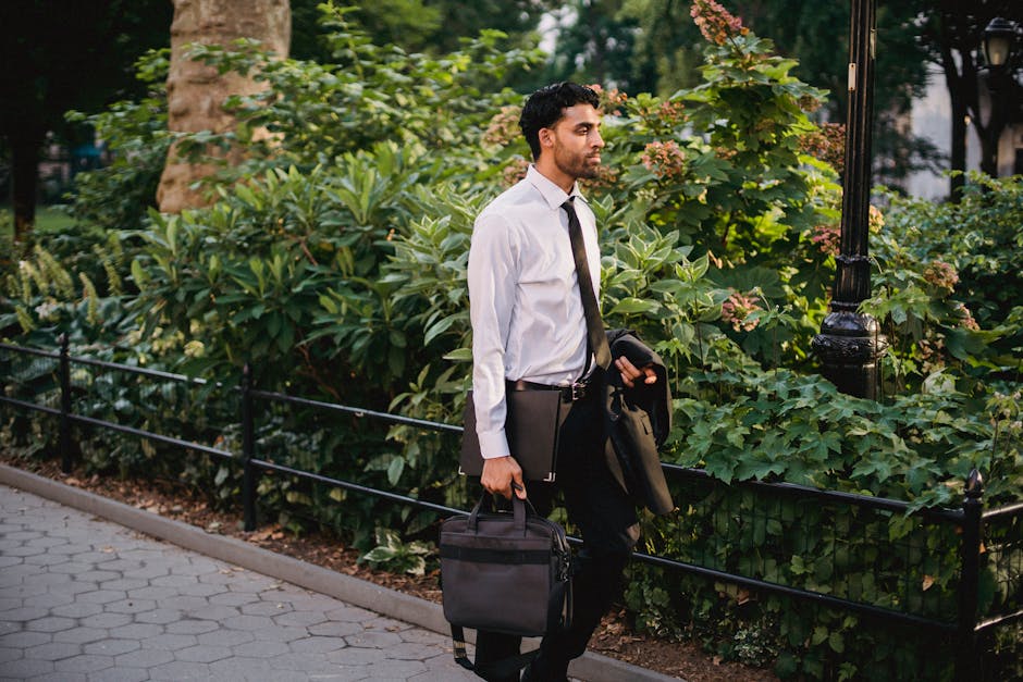 Businessman in formal attire carrying a briefcase, walking through a lush urban park on a sunny day.