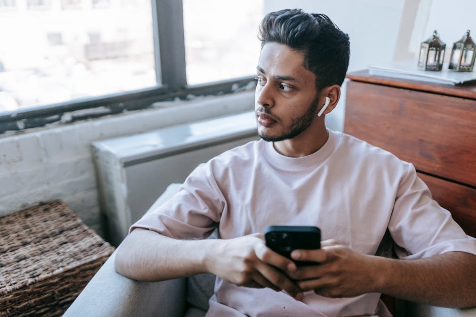 Focused bearded Indian male looking away and listening to music in wireless earphones while texting message on cellphone in armchair