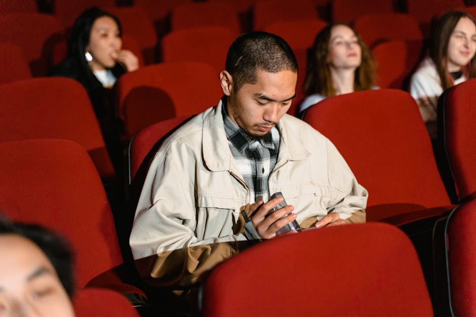 Asian man using phone in a movie theater while others watch the screen attentively.
