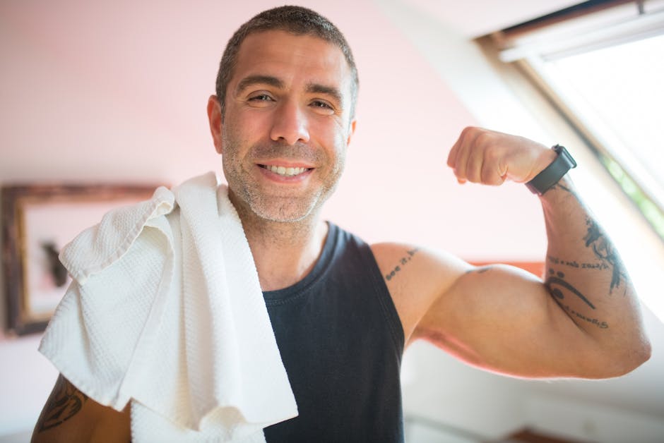 Cheerful man showing strength while flexing bicep indoors with a towel over shoulder, demonstrating fitness and positivity.