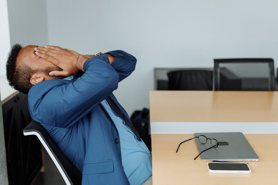 A stressed businessman in a suit covers his face while sitting at a desk with office tools.