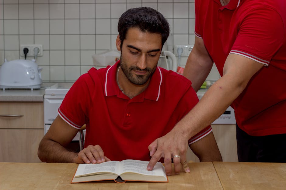 Two men in red shirts engaged in reading and discussion at a kitchen table, focusing on a book.