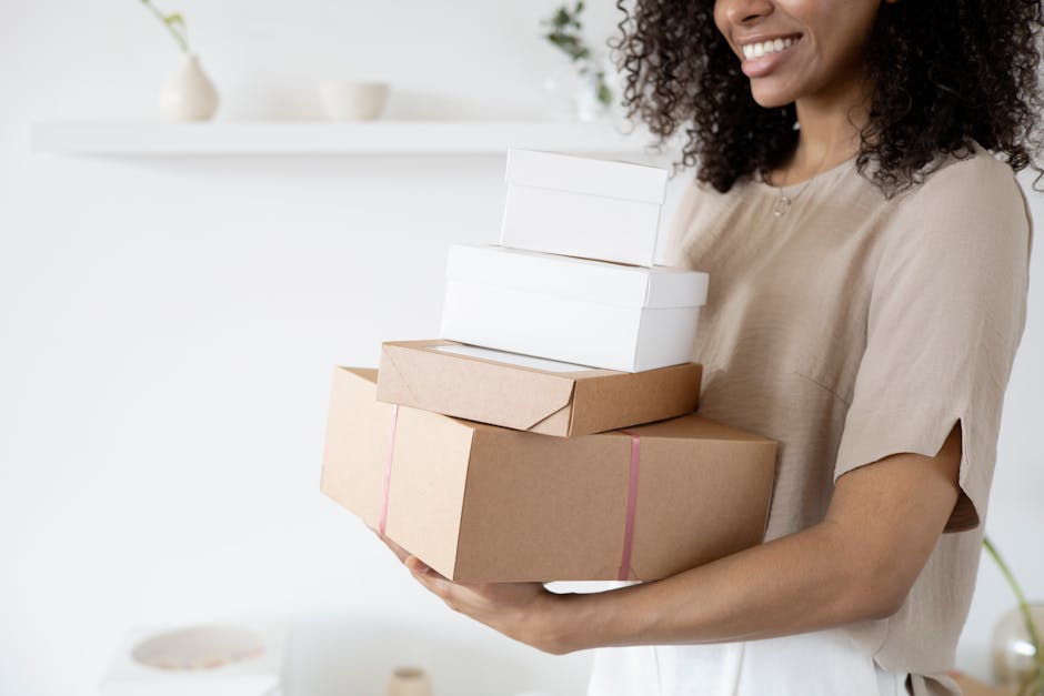 Woman with curly hair carrying stack of packaging boxes in a bright room.