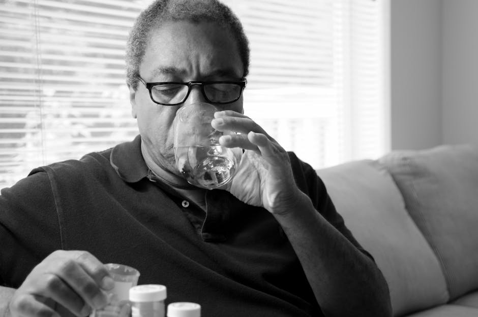 A man sits on a couch taking pills with a glass of water, suggesting self-care or medication.