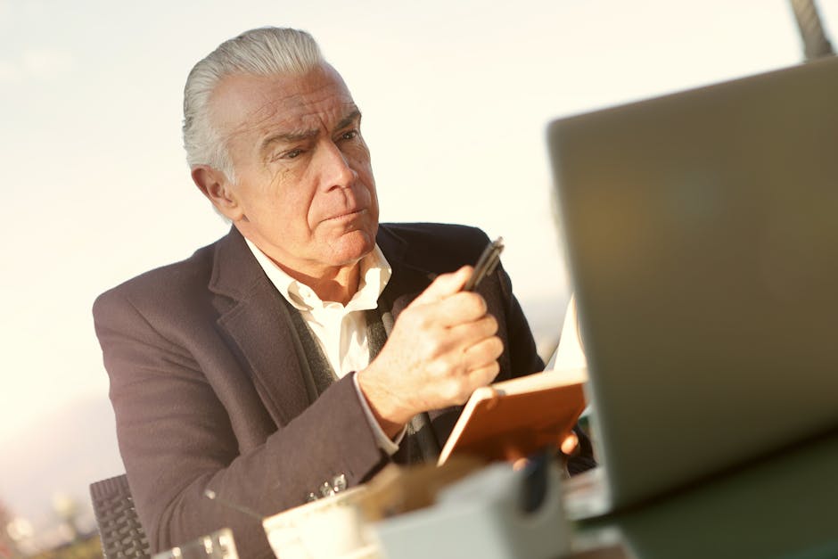 Senior man working at a desk with a laptop, notebook, and coffee, showing concentration.