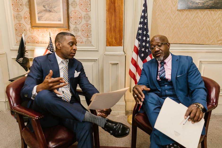 Two African American men in formal attire discussing documents in an office setting with flags in the background.