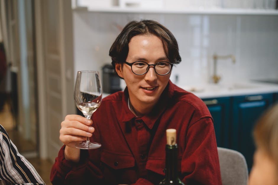 An Asian man in glasses enjoying a glass of wine indoors, sharing a warm moment with friends.
